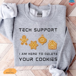 May include: A light gray crewneck sweatshirt featuring the black text TECH SUPPORT and I AM HERE TO DELETE YOUR COOKIES. Between the lines of text are four brown gingerbread-style cookies shaped like a person, a star, an ornament, and a house.