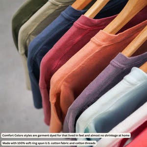 May include: A collection of crew neck t-shirts in various colors including olive, navy, red, orange, purple, and light blue, hanging on wooden hangers. The shirts feature a garment-dyed finish and are made with 100% soft ring spun U.S. cotton fabric.