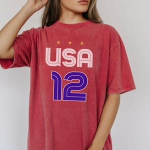 K&ouml;nnte beinhalten: Rotes T-Shirt mit dem Wort "USA" in Rosa und Wei&szlig; und der Zahl "12" in Blau und Lila. Das T-Shirt hat einen Vintage-Look. &Uuml;ber dem Wort "USA" befinden sich drei goldene Sterne.