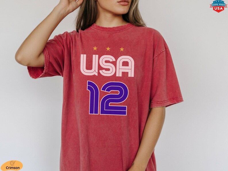 K&ouml;nnte beinhalten: Rotes T-Shirt mit dem Wort "USA" in Rosa und Wei&szlig; und der Zahl "12" in Blau und Lila. Das T-Shirt hat einen Vintage-Look. &Uuml;ber dem Wort "USA" befinden sich drei goldene Sterne.