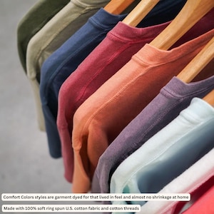 K&ouml;nnte beinhalten: Eine Reihe farbenfroher Rundhals-T-Shirts in Gr&uuml;n-, Blau-, Rot- und Oranget&ouml;nen, die auf Holzb&uuml;geln h&auml;ngen. Die Shirts bestehen aus weichem Baumwollstoff. Der Text auf dem Bild besagt, dass die Shirts st&uuml;ckgef&auml;rbt sind und aus weicher Ringspinnbaumwolle hergestellt werden.