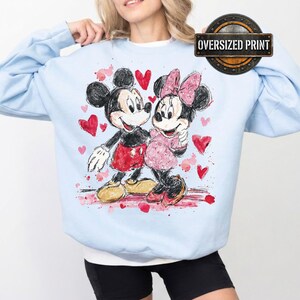 Op de afbeelding: Lichtblauwe sweatshirt met een aquarel illustratie van Mickey en Minnie Mouse, omringd door rode harten. Het ontwerp bevat de tekst "Oversized Print" in een cirkelvormig embleem.