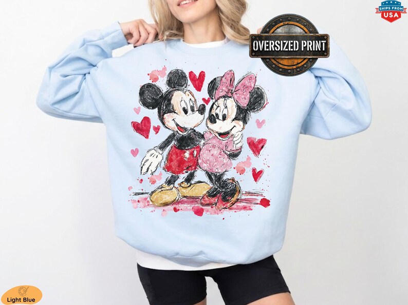 Op de afbeelding: Lichtblauwe sweatshirt met een aquarel illustratie van Mickey en Minnie Mouse, omringd door rode harten. Het ontwerp bevat de tekst "Oversized Print" in een cirkelvormig embleem.