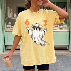May include: A butter yellow short-sleeve t-shirt featuring a graphic of a white ghost wearing a cowboy hat. The numbers 6 and 7 are printed in orange on either side of the ghost. The shirt has a crew neckline and a loose, relaxed fit.