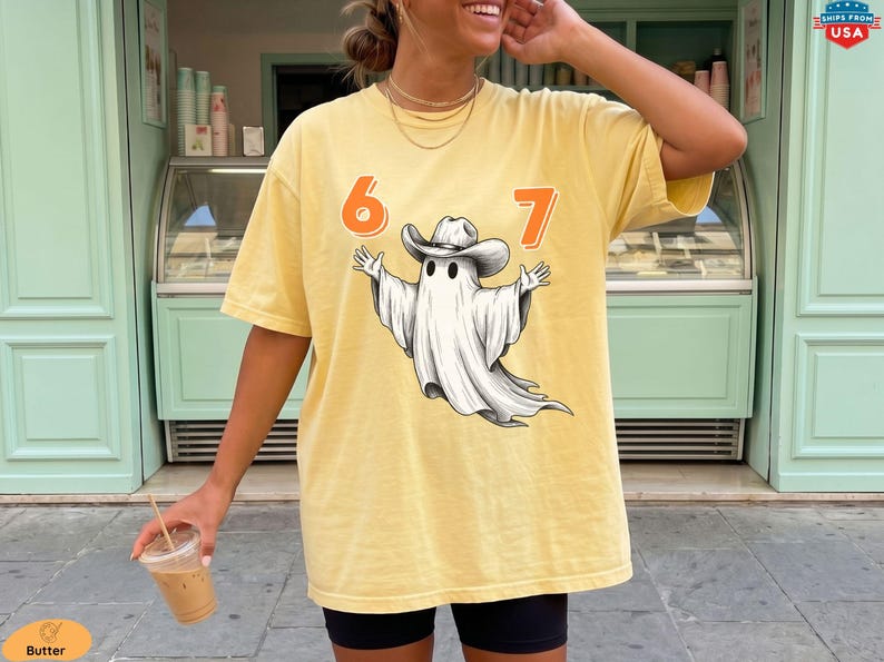 May include: A butter yellow short-sleeve t-shirt featuring a graphic of a white ghost wearing a cowboy hat. The numbers 6 and 7 are printed in orange on either side of the ghost. The shirt has a crew neckline and a loose, relaxed fit.