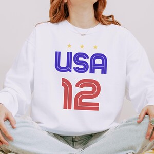 K&ouml;nnte beinhalten: Wei&szlig;es Langarm-Sweatshirt mit dem Wort "USA" in Blau und Rot und der Zahl "12" in Rot. &Uuml;ber dem Wort "USA" befinden sich drei gelbe Sterne.