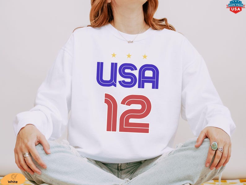 K&ouml;nnte beinhalten: Wei&szlig;es Langarm-Sweatshirt mit dem Wort "USA" in Blau und Rot und der Zahl "12" in Rot. &Uuml;ber dem Wort "USA" befinden sich drei gelbe Sterne.