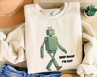 May include: Cream-colored sweatshirt featuring a green robot illustration with the text "Beep Boop I'm Out." The robot is depicted walking. The sweatshirt is paired with blue denim shorts and a brown hat.