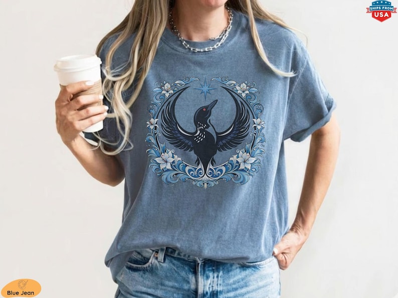May include: Blue jean t-shirt featuring a detailed illustration of a black bird with outstretched wings, surrounded by a blue floral wreath and a star. The bird has a red eye. The shirt is a faded blue color.