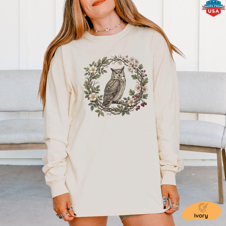 May include: Ivory long-sleeve shirt featuring a detailed illustration of an owl perched within a floral wreath. The owl is depicted in shades of brown and gray, surrounded by green leaves, white flowers, and red berries. The shirt has a relaxed fit.