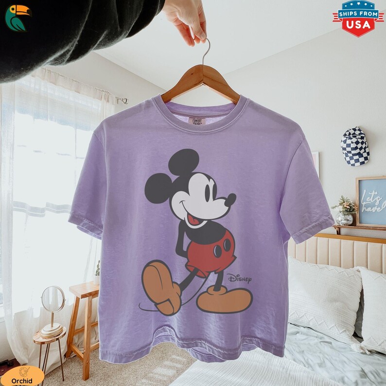 K&ouml;nnte beinhalten: Ein lila T-Shirt mit einer gro&szlig;en Mickey Mouse-Grafik. Die Zeichentrickfigur ist in ihrer klassischen Pose dargestellt, mit schwarzen Ohren, einem wei&szlig;en Gesicht, roten Shorts und braunen Schuhen. Das Disney-Logo ist unten rechts aufgedruckt.