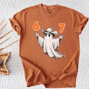 May include: A rust-colored short-sleeve t-shirt featuring a graphic of a white ghost wearing a cowboy hat with its arms outstretched. The numbers 6 and 7 are printed in orange above the ghost. The shirt has a crew neckline and a relaxed fit.