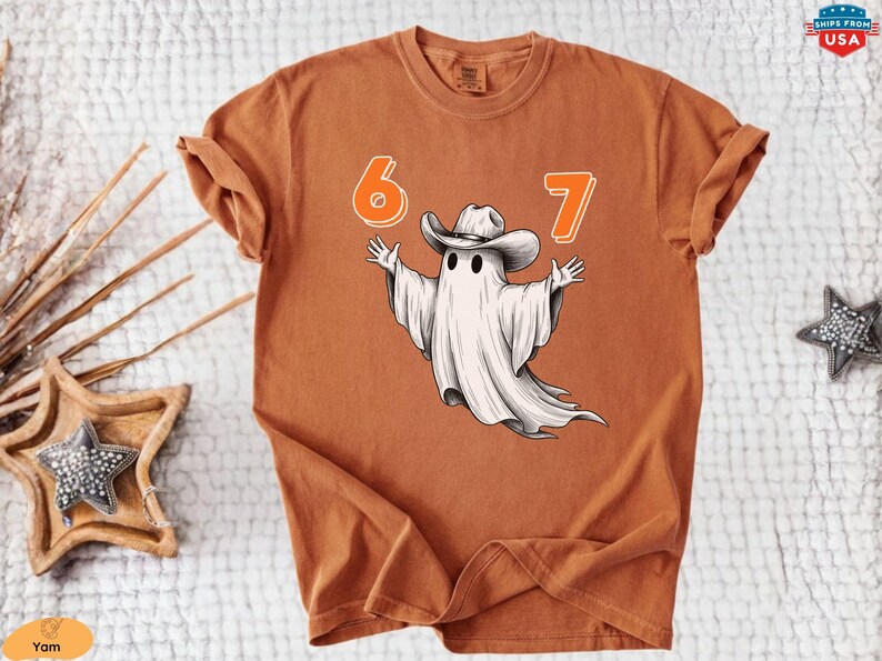 May include: A rust-colored short-sleeve t-shirt featuring a graphic of a white ghost wearing a cowboy hat with its arms outstretched. The numbers 6 and 7 are printed in orange above the ghost. The shirt has a crew neckline and a relaxed fit.