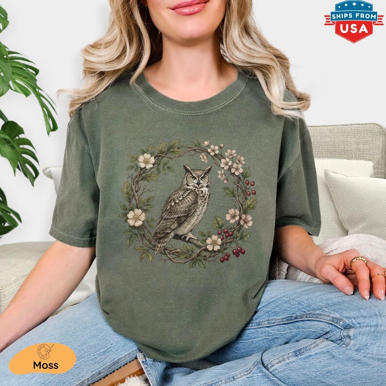 May include: Moss green t-shirt featuring a detailed illustration of an owl perched on a branch within a floral wreath. The design includes white flowers and red berries. The shirt has a classic crew neck and short sleeves.