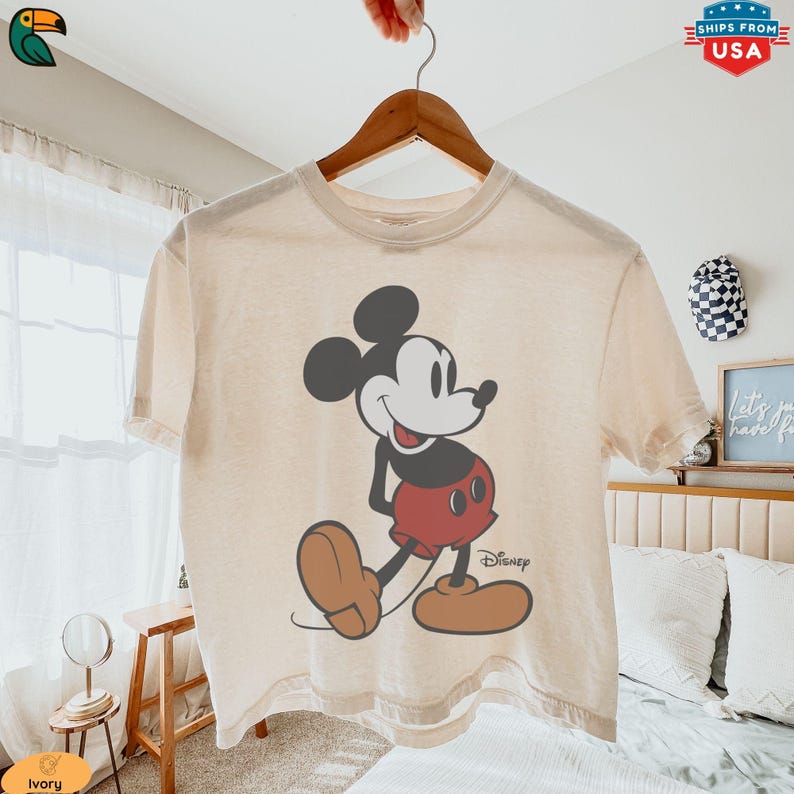 K&ouml;nnte beinhalten: Cremefarbenes T-Shirt mit einem klassischen Mickey Mouse-Motiv. Die Zeichentrickfigur ist in Schwarz, Rot und Beige dargestellt. Das Disney-Logo ist darunter aufgedruckt. Das Shirt h&auml;ngt an einem Holzb&uuml;gel.