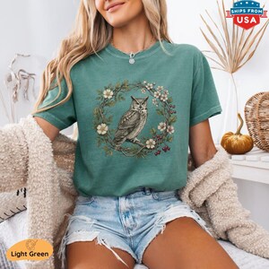 May include: Light green t-shirt featuring a detailed illustration of an owl perched on a branch within a floral wreath. The design includes white flowers and red berries. The shirt is a solid light green color.