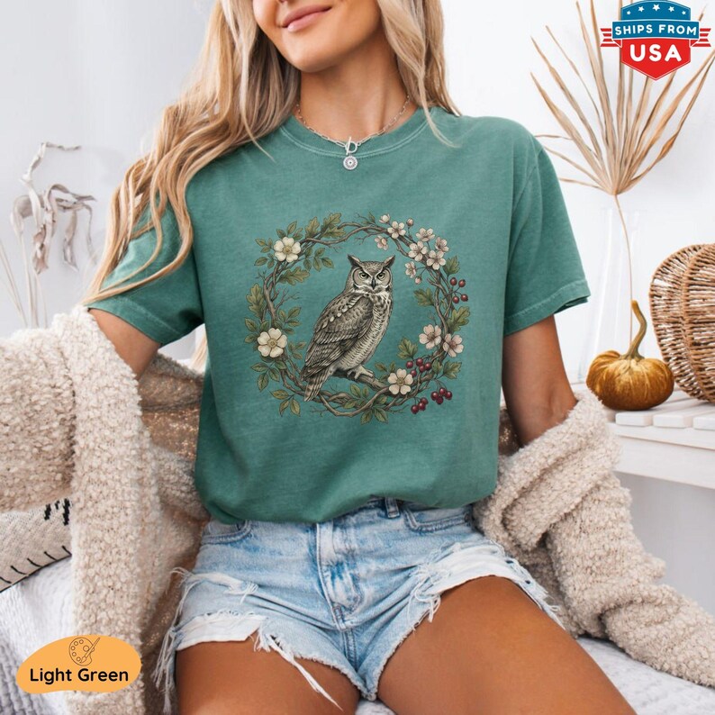 May include: Light green t-shirt featuring a detailed illustration of an owl perched on a branch within a floral wreath. The design includes white flowers and red berries. The shirt is a solid light green color.