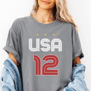 K&ouml;nnte beinhalten: Graues T-Shirt mit dem Wort "USA" in Wei&szlig; und Rot und der Zahl "12" in Rot. &Uuml;ber dem Wort "USA" befinden sich drei goldene Sterne. Das Shirt ist aus einem weichen Material.
