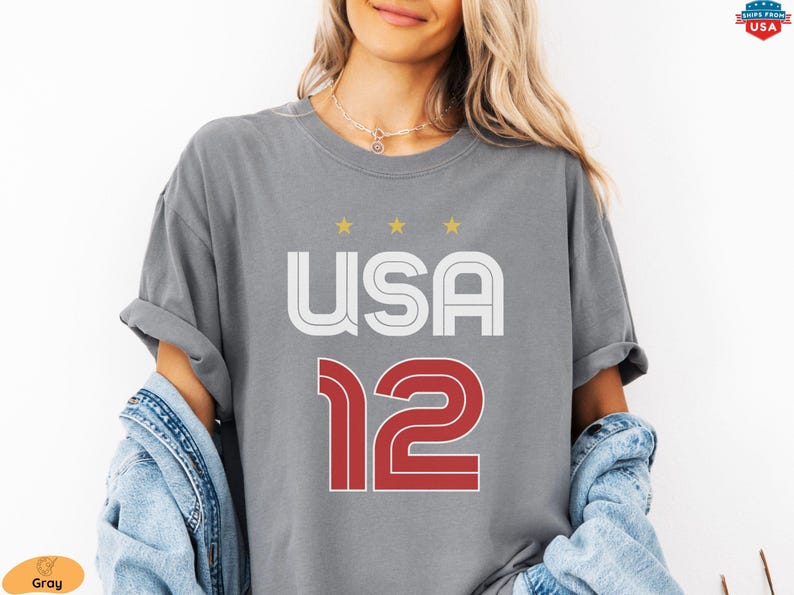 K&ouml;nnte beinhalten: Graues T-Shirt mit dem Wort "USA" in Wei&szlig; und Rot und der Zahl "12" in Rot. &Uuml;ber dem Wort "USA" befinden sich drei goldene Sterne. Das Shirt ist aus einem weichen Material.