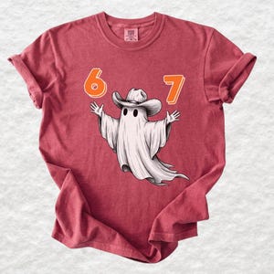 May include: A crimson short-sleeve t-shirt featuring a graphic of a white ghost wearing a cowboy hat with its arms outstretched. The numbers 6 and 7 are printed in orange above the ghost. The shirt has a crew neckline and a relaxed fit.