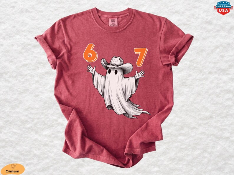 May include: A crimson short-sleeve t-shirt featuring a graphic of a white ghost wearing a cowboy hat with its arms outstretched. The numbers 6 and 7 are printed in orange above the ghost. The shirt has a crew neckline and a relaxed fit.