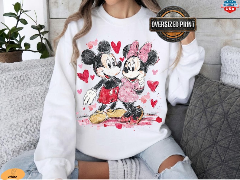 May include: White sweatshirt featuring a watercolor illustration of Mickey and Minnie Mouse embracing, surrounded by red hearts. Mickey wears red shorts, and Minnie wears a pink dress and bow. The sweatshirt has an "Oversized Print" label.