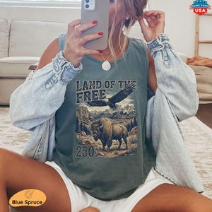 May include: A sage green tank top with a graphic featuring an eagle, bison, mountains, and the text "LAND OF THE FREE" and "250". The tank top is worn with a light blue denim jacket and white shorts. The image also includes a phone and jewelry.