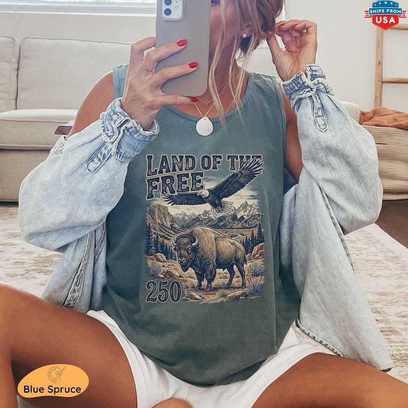 May include: A sage green tank top with a graphic featuring an eagle, bison, mountains, and the text "LAND OF THE FREE" and "250". The tank top is worn with a light blue denim jacket and white shorts. The image also includes a phone and jewelry.