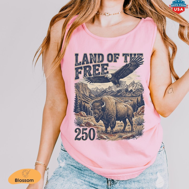 May include: Pink tank top with a graphic design featuring the text "LAND OF THE FREE", a soaring eagle, and a bison in a mountain landscape. The number "250" is below the bison. The tank top is sleeveless.