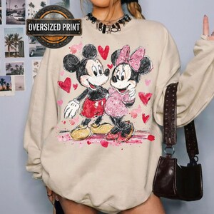 Op de afbeelding: Zandkleurige oversized print sweatshirt met Mickey en Minnie Mouse die elkaar omarmen, omringd door roze harten. Het grafische ontwerp bevat de tekst "Oversized Print" in een cirkelvormig embleem. Een donkerbruine handtas hangt aan de schouder.