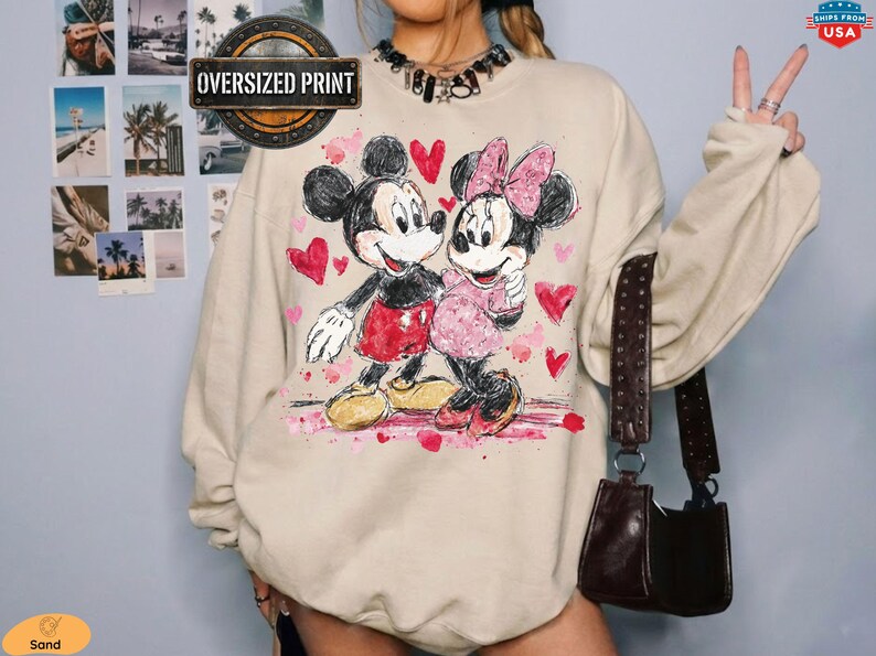 Op de afbeelding: Zandkleurige oversized print sweatshirt met Mickey en Minnie Mouse die elkaar omarmen, omringd door roze harten. Het grafische ontwerp bevat de tekst "Oversized Print" in een cirkelvormig embleem. Een donkerbruine handtas hangt aan de schouder.