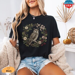 May include: Black t-shirt featuring a detailed illustration of an owl perched on a branch within a floral wreath. The design includes white flowers, green leaves, and red berries. The shirt is a classic crew neck style.