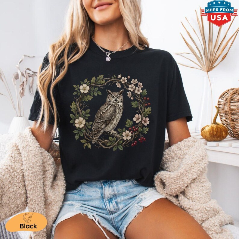 May include: Black t-shirt featuring a detailed illustration of an owl perched on a branch within a floral wreath. The design includes white flowers, green leaves, and red berries. The shirt is a classic crew neck style.
