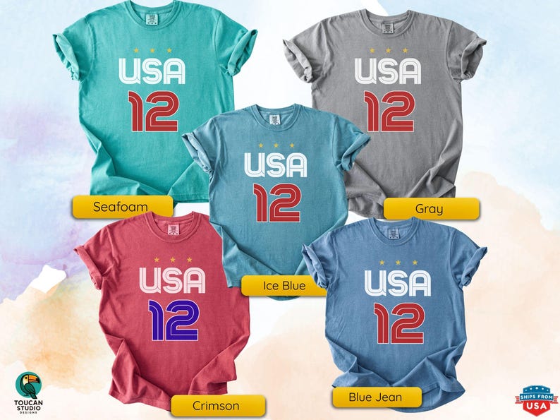 K&ouml;nnte beinhalten: F&uuml;nf T-Shirts in verschiedenen Farben: Seegr&uuml;n, Grau, Eisblau, Karmesinrot und Jeansblau. Jedes Shirt zeigt den Text "USA 12" in Wei&szlig; und Rot. Die Shirts sind kurz&auml;rmelig und haben einen l&auml;ssigen Stil.