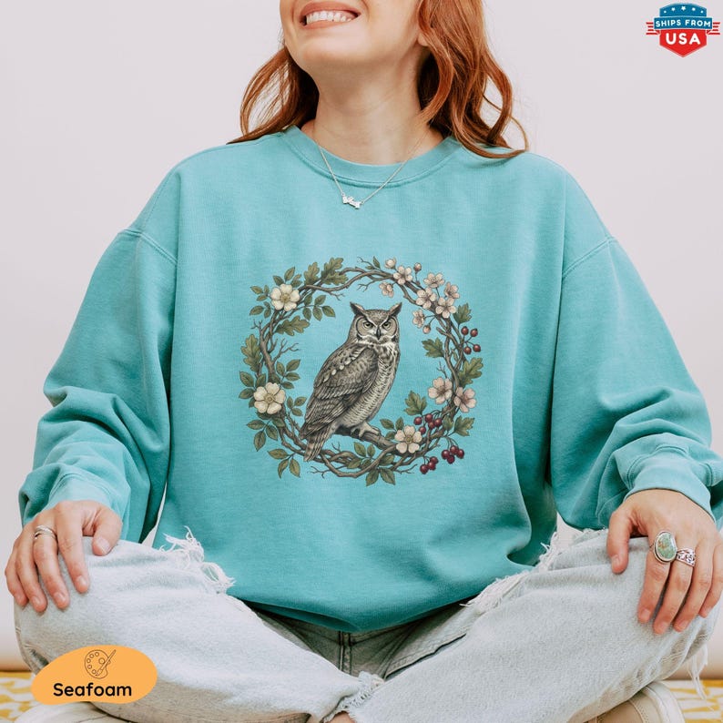 May include: Seafoam green sweatshirt featuring a detailed illustration of an owl perched on a branch within a floral wreath. The owl is depicted in shades of gray and brown, surrounded by white flowers and red berries. The sweatshirt has a relaxed fit.