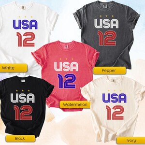 K&ouml;nnte beinhalten: F&uuml;nf T-Shirts in verschiedenen Farben: Wei&szlig;, Pfeffer, Wassermelone, Schwarz und Elfenbein. Jedes Shirt zeigt den Text "USA 12" in einer kr&auml;ftigen, stilisierten Schrift mit drei Sternen &uuml;ber "USA".