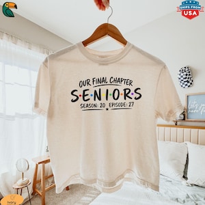 May include: An ivory short-sleeve t-shirt with black text that reads OUR FINAL CHAPTER SENIORS SEASON: 20 EPISODE: 27. The word SENIORS features colorful dots between the letters, mimicking a television show logo style on the front of the shirt.