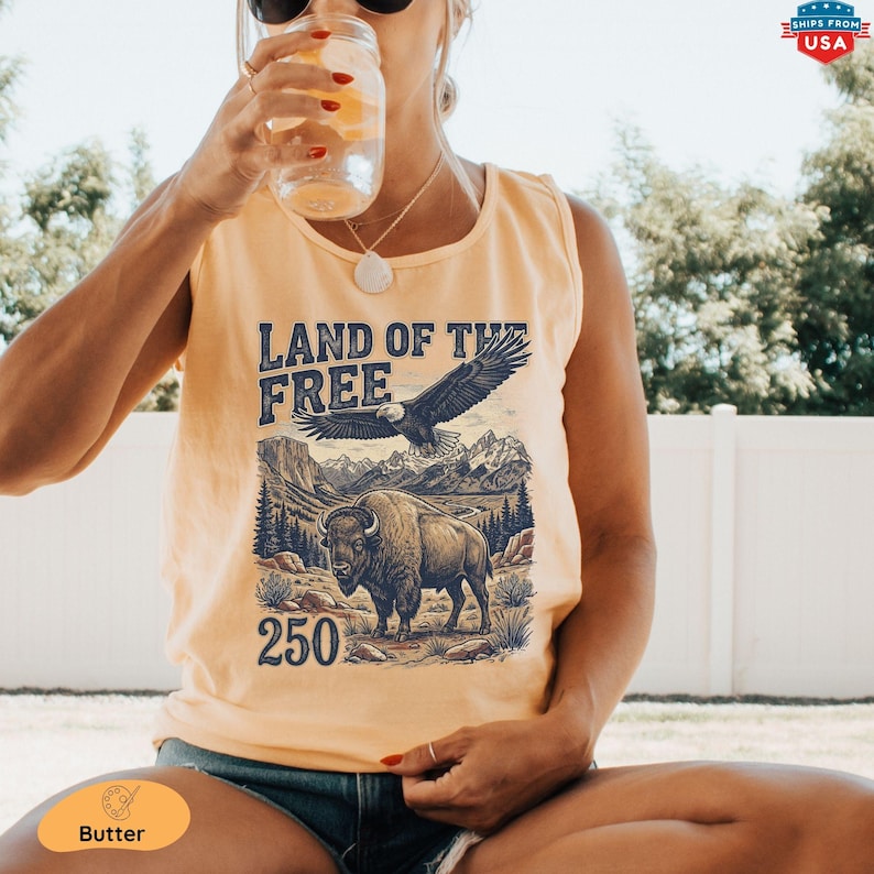 A woman sitting outdoors wearing a sleeveless graphic tank top and holding a jar with a cold beverage. The shirt features a vintage-style retro illustration of mountains, a flying bald eagle, an American bison, and the text "LAND OF THE FREE" and "250".