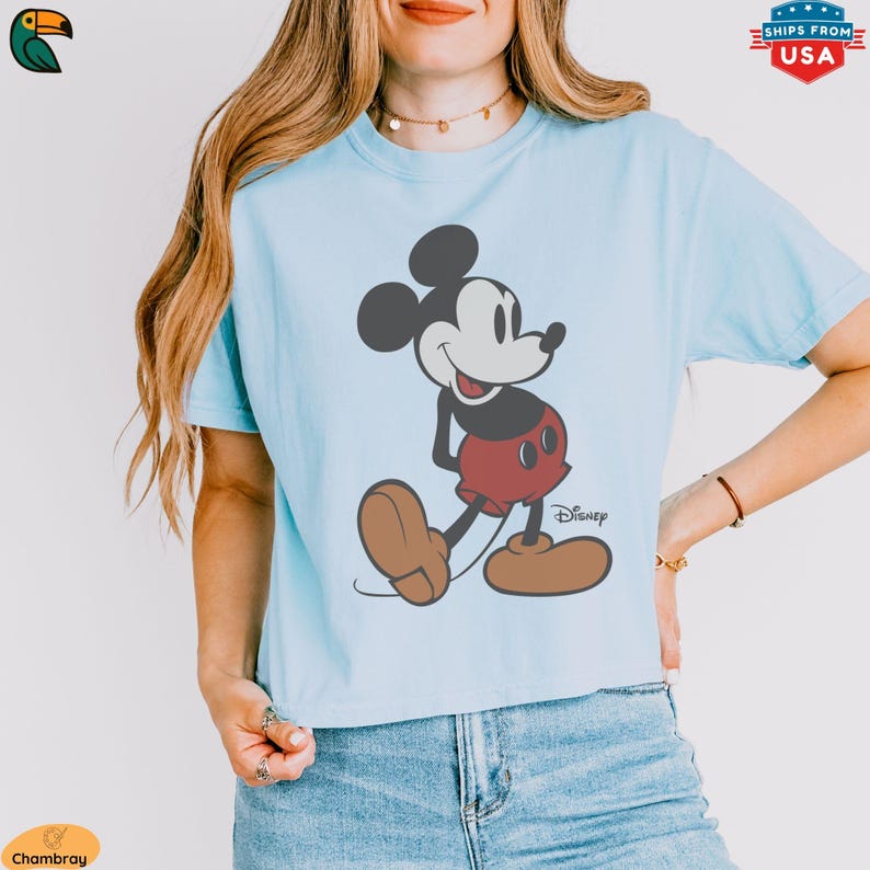 K&ouml;nnte beinhalten: Hellblaues T-Shirt mit einem Mickey Mouse-Motiv. Die Zeichentrickfigur ist in Schwarz, Rot und Braun dargestellt, mit dem Disney-Logo darunter. Das T-Shirt hat einen l&auml;ssigen Stil.