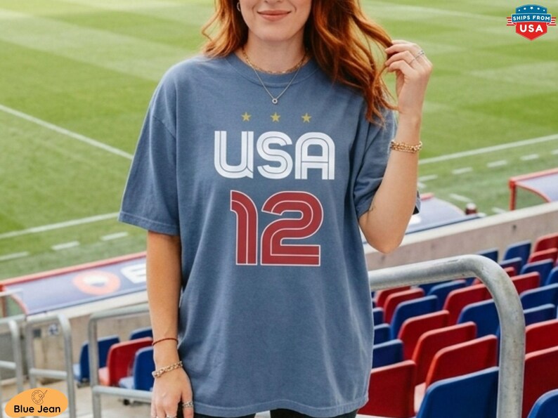 K&ouml;nnte beinhalten: Ein blaues T-Shirt mit dem Text "USA 12" in Wei&szlig; und Rot. Das Shirt hat einen Rundhalsausschnitt und kurze &Auml;rmel. Das Bild ist im Freien aufgenommen, mit einem Stadion im Hintergrund.