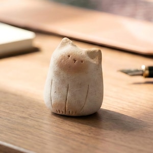 Puede incluir: Pequeña figura de gato de cerámica blanquecina, con cuerpo redondeado y cara marrón claro. El gato tiene detalles sencillos grabados y está sobre una superficie de madera. Un bolígrafo y un portátil están en el fondo.