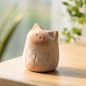 Könnte beinhalten: Kleine, handgefertigte Katzenfigur aus Keramik in hellbrauner Farbe. Die Katze hat eine einfache, abgerundete Form mit geätzten Details und einem etwas dunkleren braunen Gesicht. Sie steht auf einer Holzoberfläche, mit Grünzeug im Hintergrund.