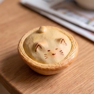 Könnte beinhalten: Kleine, runde Keramik-Schmuckdose mit einem Katzengesicht-Design auf dem Deckel. Die Dose ist hellgelb mit braunen Akzenten, und die Katzenmerkmale sind braun gezeichnet. Sie steht auf einer Holzoberfläche.