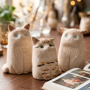 May include: Three ceramic cat figurines in varying shades of beige and tan sit on a wooden table. The cats have blue or green eyes and are in a sitting position. An open book with illustrations is in the foreground.
