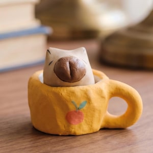Pode incluir: Uma pequena caneca de cerâmica amarela texturizada com uma fruta laranja pintada com uma folha verde na frente. Dentro da caneca está uma figura de capivara esculpida em castanho claro com um nariz castanho escuro, visível do peito para cima.