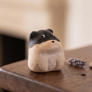 May include: A small, rounded ceramic figurine shaped like a stylized animal with a black upper head and a light beige lower body. The figure features two small ears, two white eyes with black pupils, and a tiny black nose, standing on a flat base.