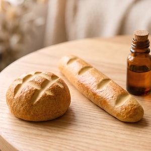 Può includere: Natura morta con un panino rotondo e una baguette dorata, accanto a una piccola bottiglia di vetro ambrato con tappo di sughero. Gli oggetti sono disposti su un tavolo di legno chiaro.