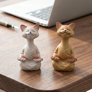 Puede incluir: Dos figuras de gato de cerámica en postura de yoga sentado. Una es blanquecina con detalles en rosa, la otra es de color canela. Ambas tienen los ojos cerrados y están sobre una superficie de madera, cerca de un portátil y un bolígrafo.