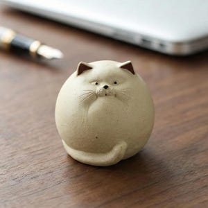 Puede incluir: Una figura de gato de cerámica beige con cuerpo redondo, orejas pequeñas y cola enroscada. El gato tiene una expresión ligeramente gruñona y está sobre una superficie de madera. Un bolígrafo y un portátil están en el fondo.