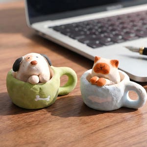 Op de afbeelding: Twee keramische beeldjes: een hond in een groene mok en een kat in een blauwe mok. De hond heeft zwarte oren en een bruine vlek. De kat is oranje en wit. De mokken staan op een houten oppervlak.